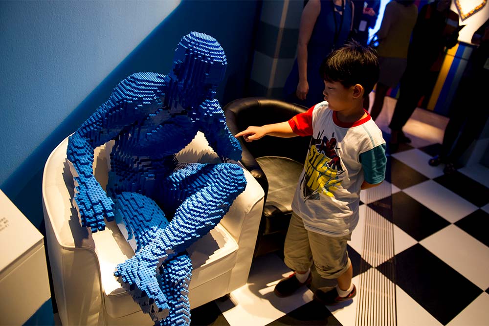 a boy touching a lego sculpture of seated man by Nathan Sawaya